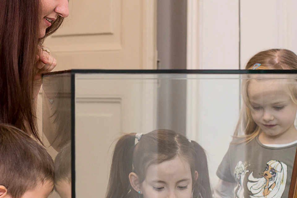 A woman and three children are looking at an ornate chair displayed in a glass case in a museum-like setting.