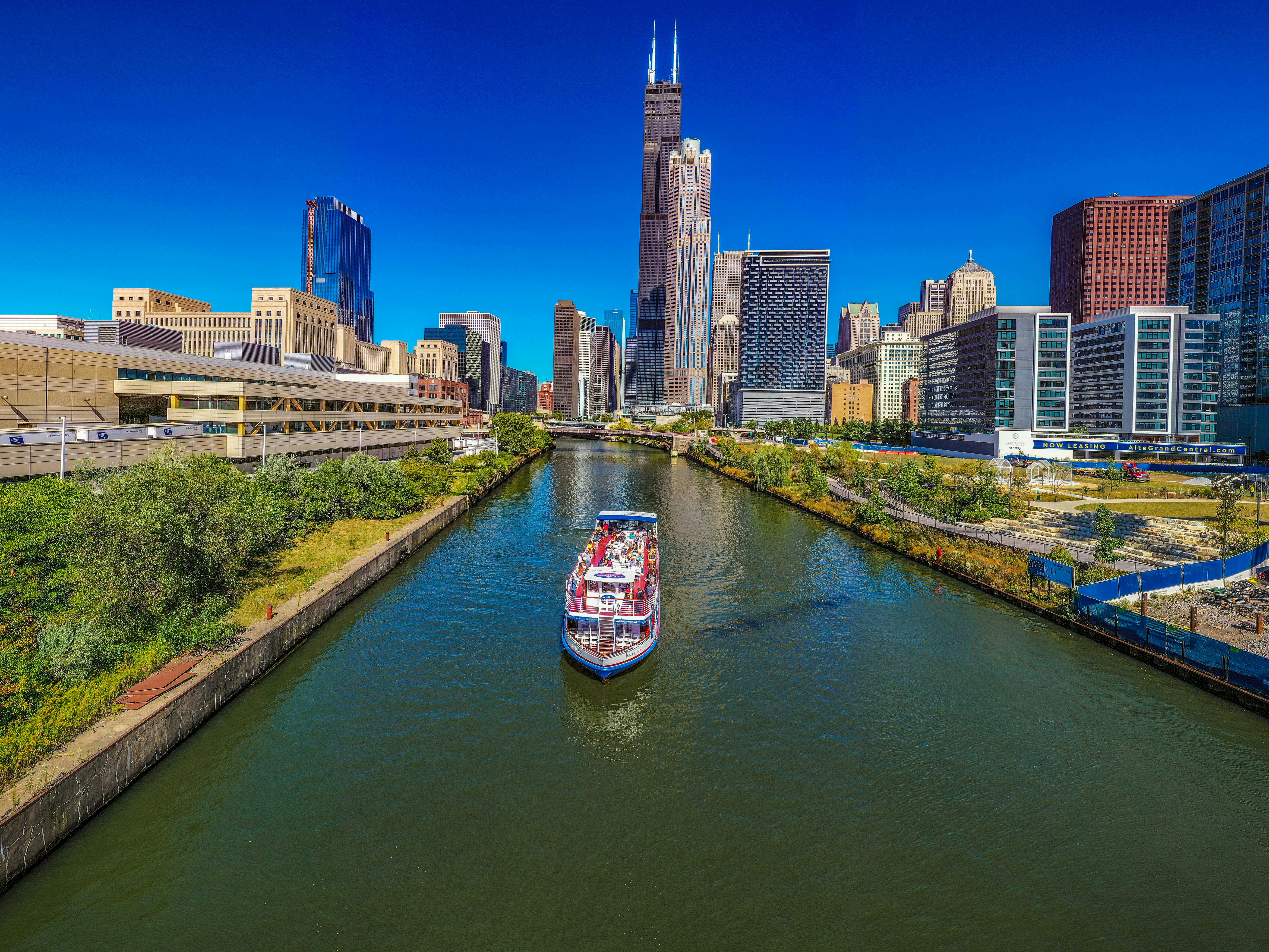 A riverboat cruises down a river flanked by urban buildings and greenery under a clear blue sky.