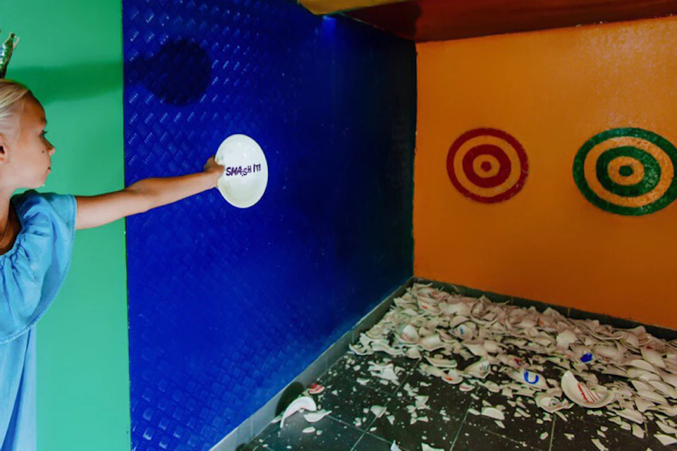A person in a blue shirt prepares to smash a white plate in a colorful room filled with broken dishes.