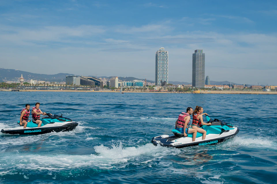 Three people ride two jet skis on a calm sea, with a cityscape and several high-rise buildings in the background.