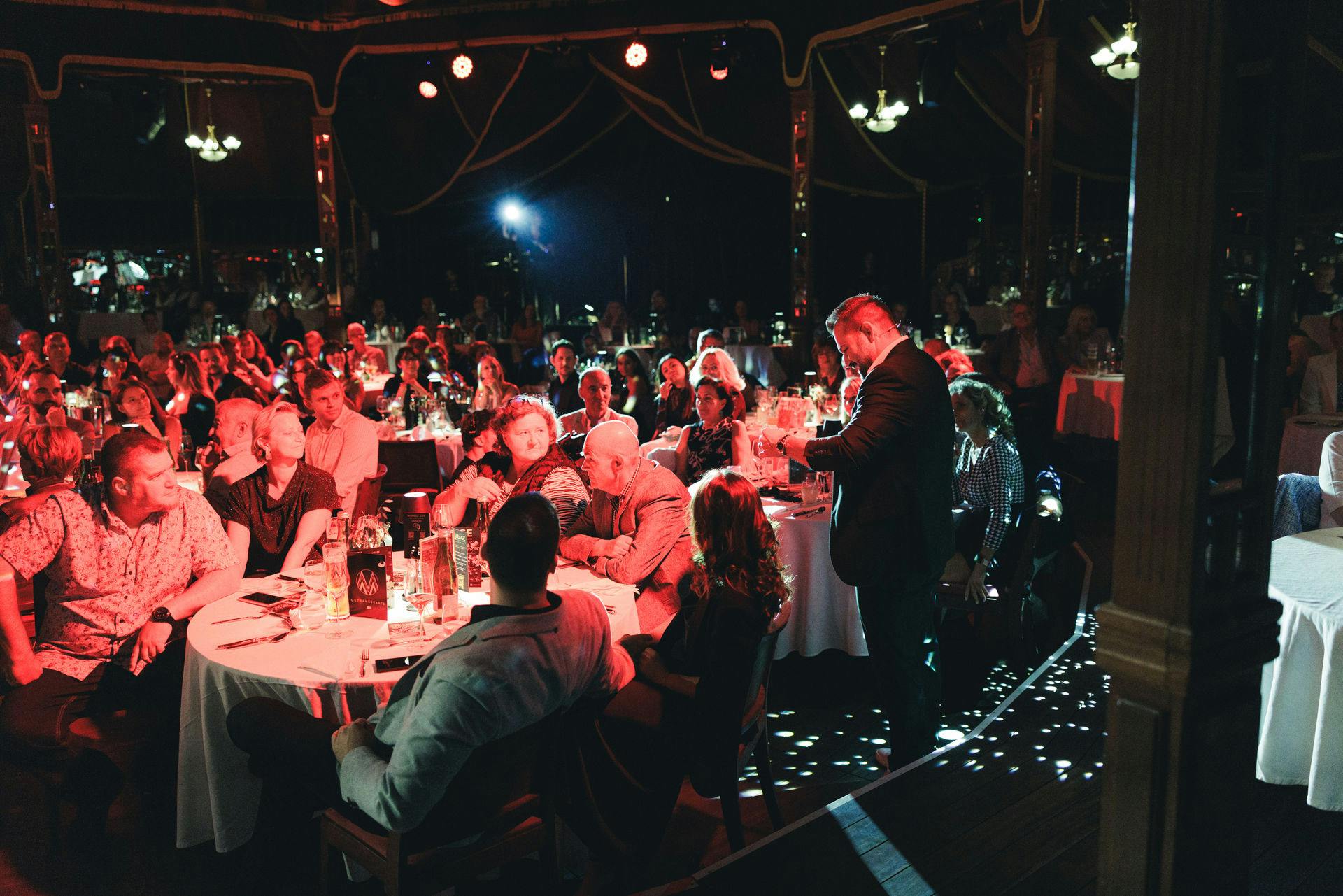 在灯光昏暗的场地，一位穿着西装的演讲者在圆桌会议上向坐着的观众讲话，并配备了红色灯光和聚光灯