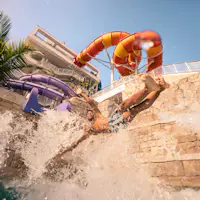 En person, der plasker i en pool fra en rutsjebane, med farverige vandrutsjebaner og en bygning i baggrunden under en klar himmel.
