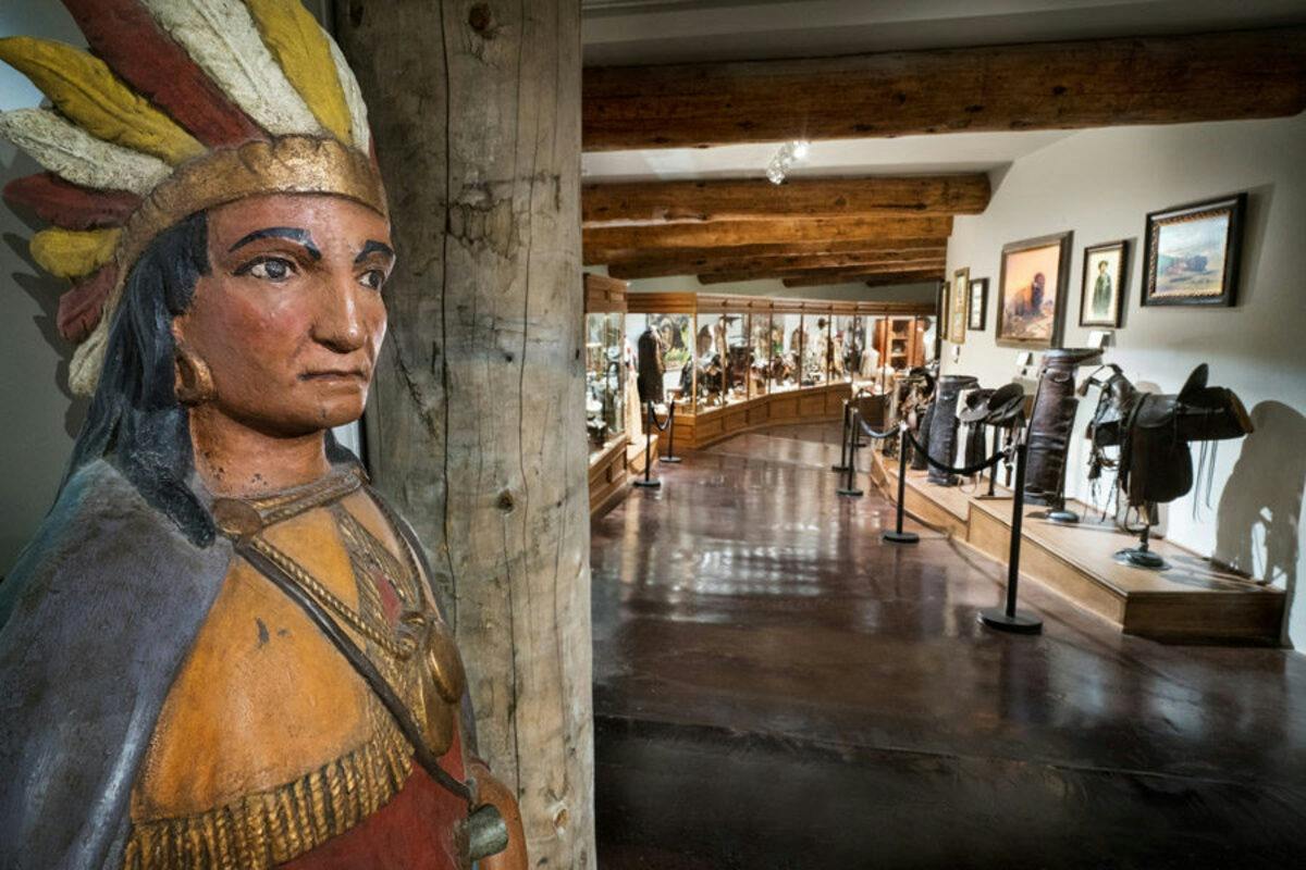 A wooden statue of a Native American in a museum, with display cases and artifacts in the background.