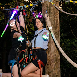 Two people wearing harnesses and glowing accessories are engaged in a rope course activity in a wooded area at dusk.