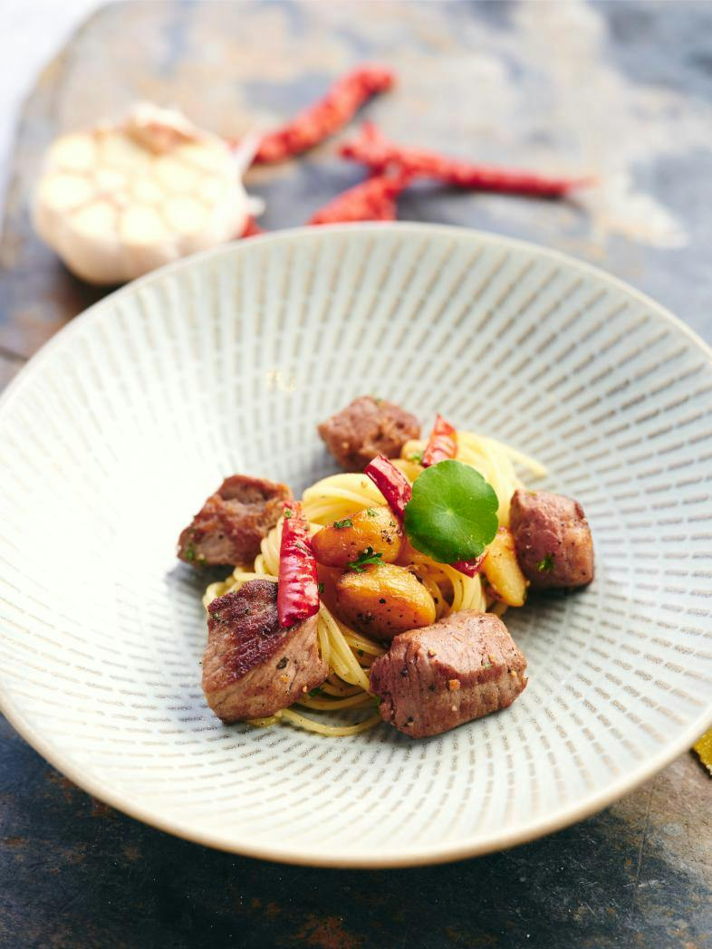 A plated dish of pasta with grilled meat, roasted garlic, and red chilies, garnished with herbs in a patterned bowl.