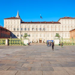 En historisk byggnad med utsmyckade portar, statyer av män till häst och människor som går på ett rymligt torg. Klarblå himmel ovanför.