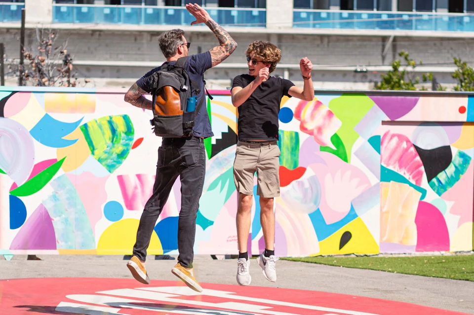 Two people high-fiving mid-air in front of a colorful mural. One has tattoos, the other is wearing sunglasses.
