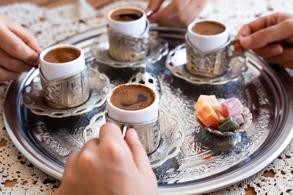 Four ornate cups of coffee on a silver tray with a small bowl of colorful sweets; hands are holding the cups.