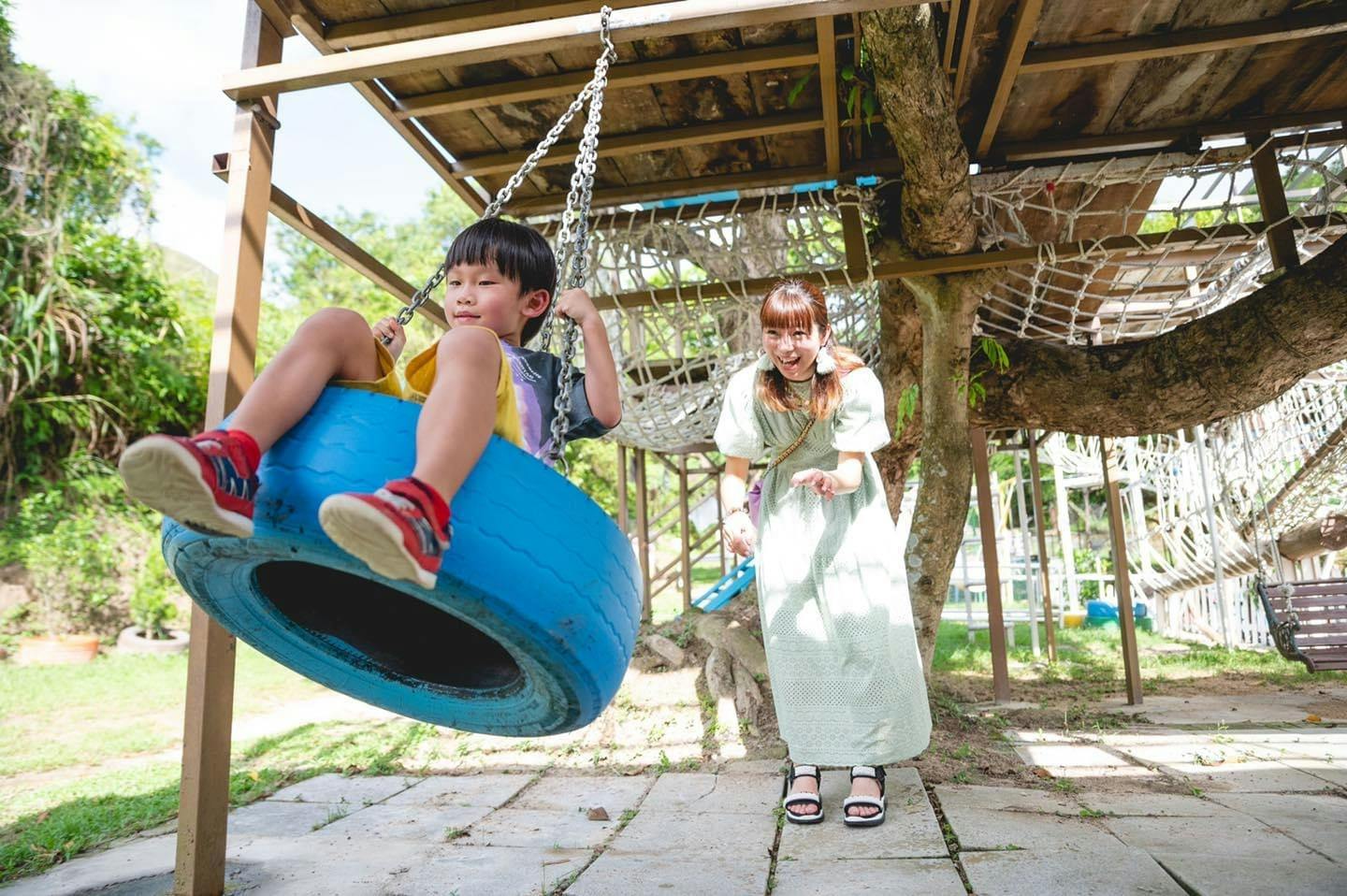 A child swings on a blue tire swing while a woman stands nearby smiling under a wooden playground structure.