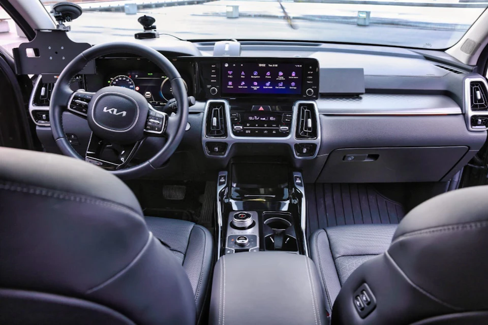 Interior of a car showing the driver's seat, steering wheel with Kia logo, dashboard with digital displays, and central console.