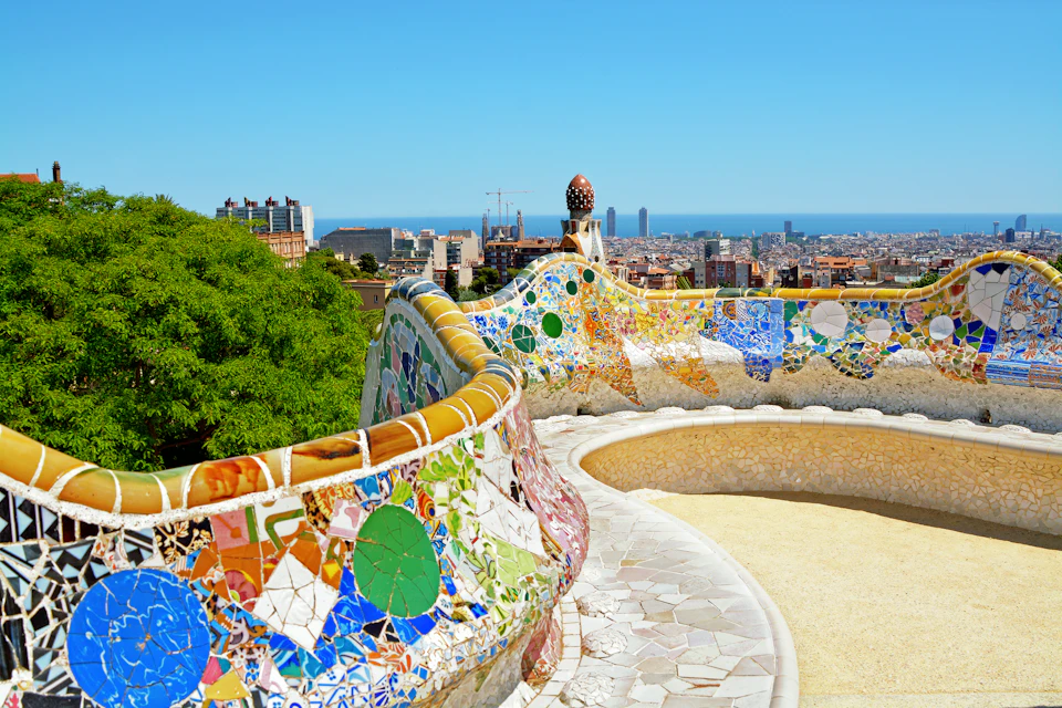 Colorful mosaic benches curve around a terrace in a park, with a view of a cityscape and blue sky in the background.