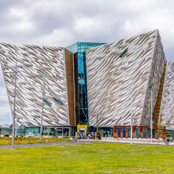Vinkelformad modern byggnad med strukturerad fasad och central ingång i glas, omgiven av grön gräsmatta och människor som går.