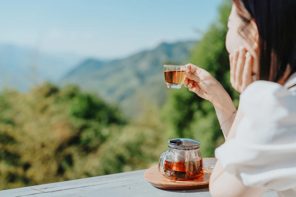 A person holds a glass of tea, with a teapot on a tray, overlooking a scenic view of hills and greenery.