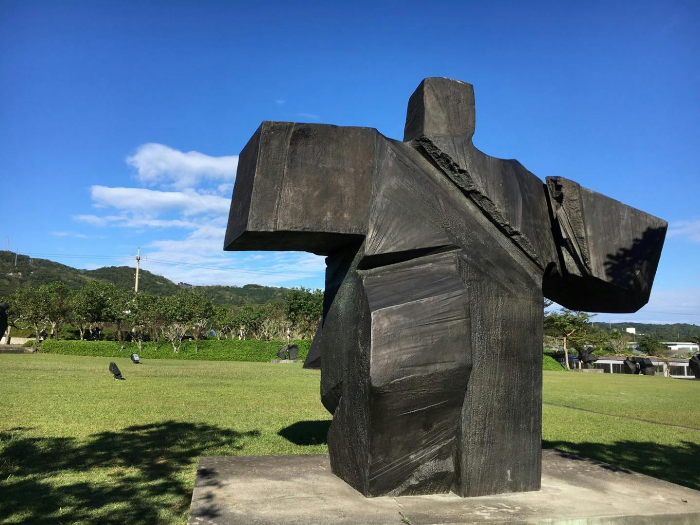 Estatua abstracta negra en un parque con césped bajo un cielo azul claro, con árboles y colinas al fondo.