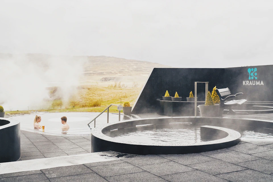 Person sitting in a steaming outdoor hot tub with a mountainous landscape in the background and modern design elements around.