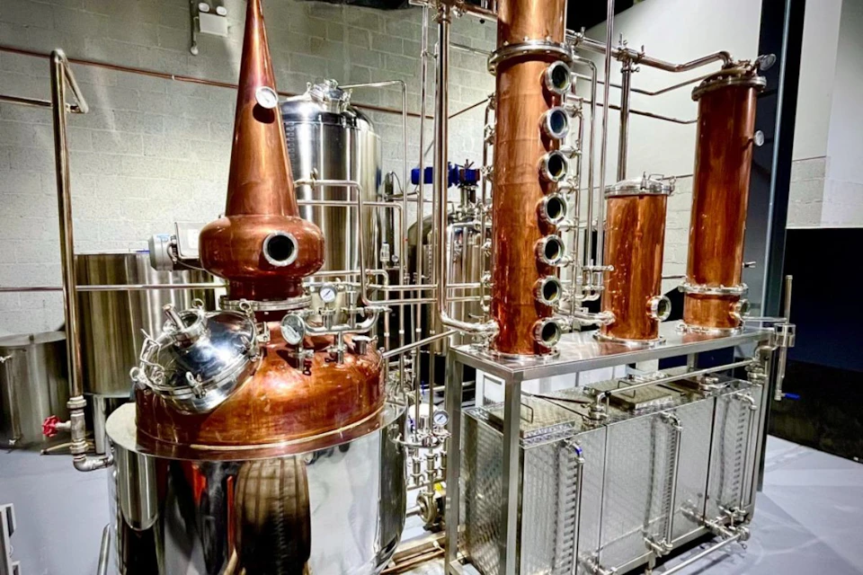 A distillery setup with copper columns, stainless steel vessels, and various pipes and gauges in an industrial space.
