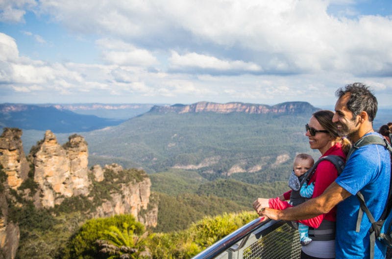 A couple with a baby in carriers stand at a scenic overlook, enjoying a view of a vast, mountainous landscape under a partly cloudy sky.
