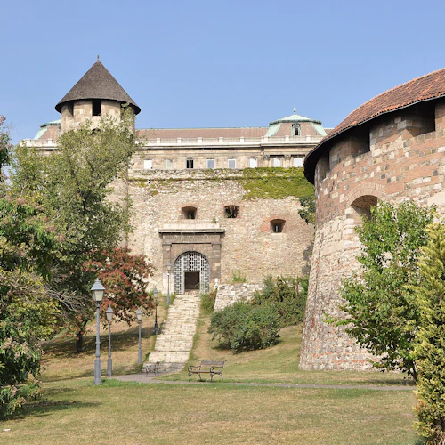 Stenfästning med runt torn, välvd ingångsport och omgivande grönska under en klarblå himmel. Bänkar och lyktstolpar framför.