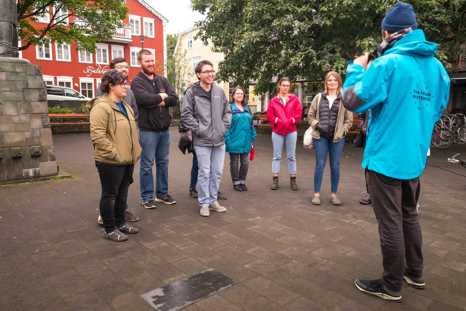 A group of people standing in an outdoor area, listening and smiling. Trees and colorful buildings are visible in the background.