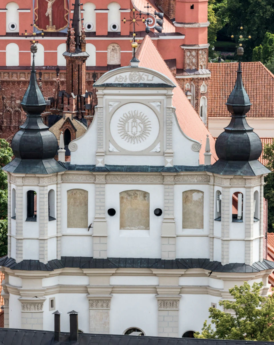 White Baroque-style church tower with two black domes, decorative details, and surrounding red-roofed buildings in the background.