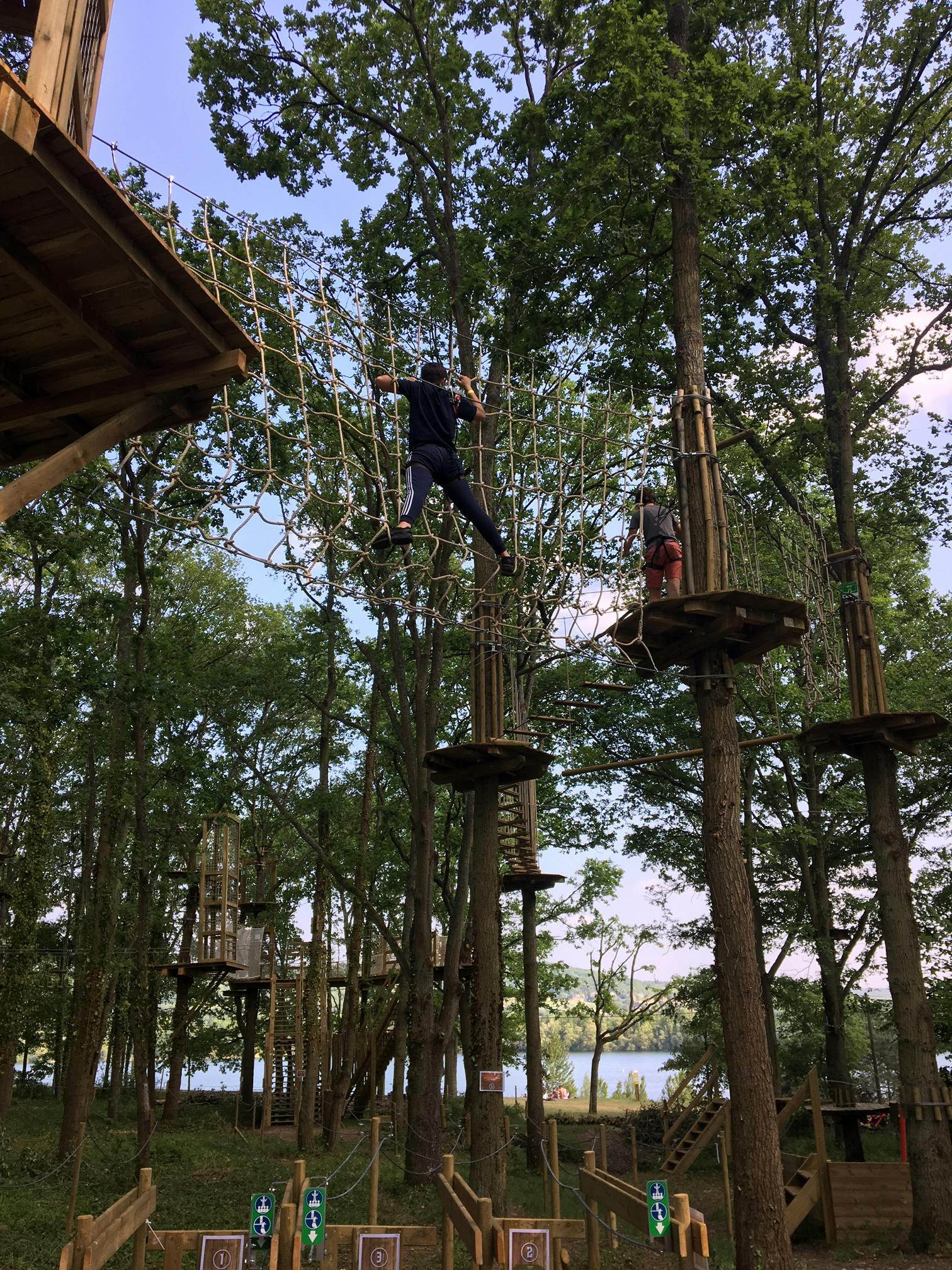 Mensen navigeren over verhoogde touwbanen en platforms in een bebost gebied met een glimp van water op de achtergrond.