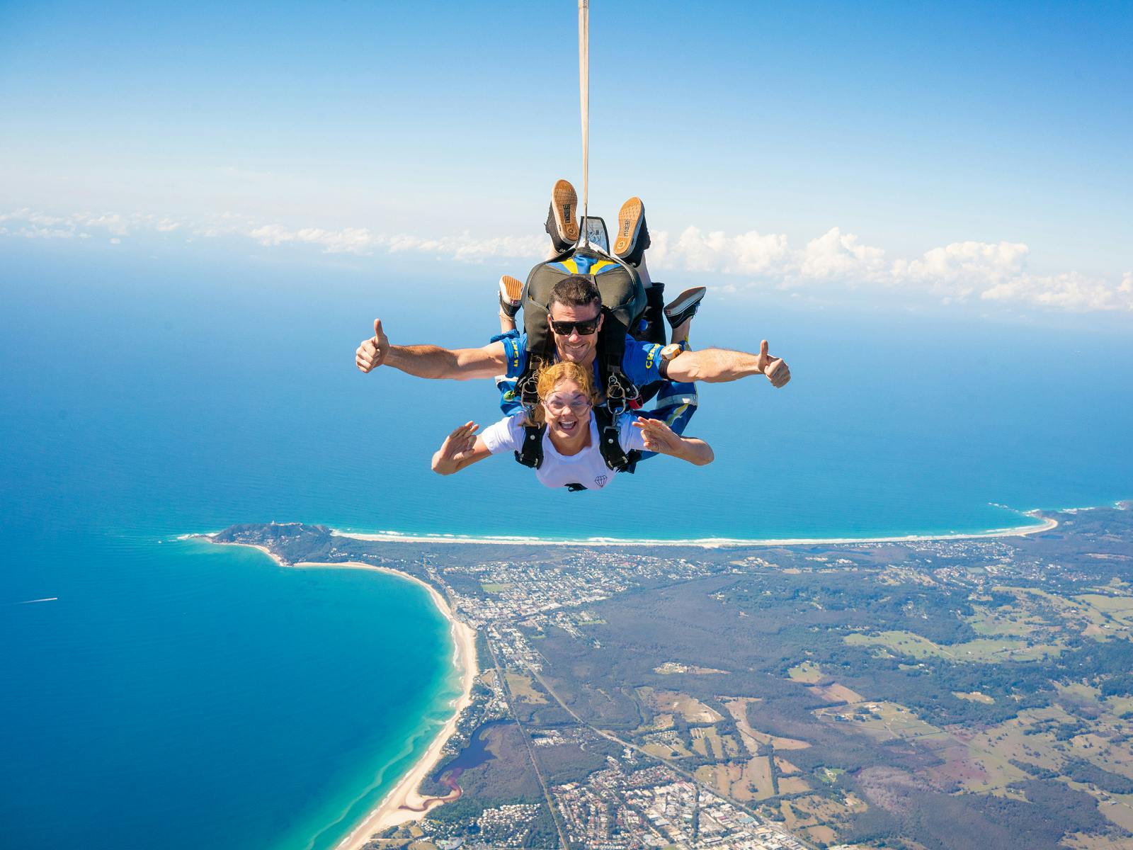 Dva lidé tandemově skáčou nad pobřežní krajinou, usmívají se a dávají palec nahoru, s oceánem, pobřežím a zemí viditelnou níže.