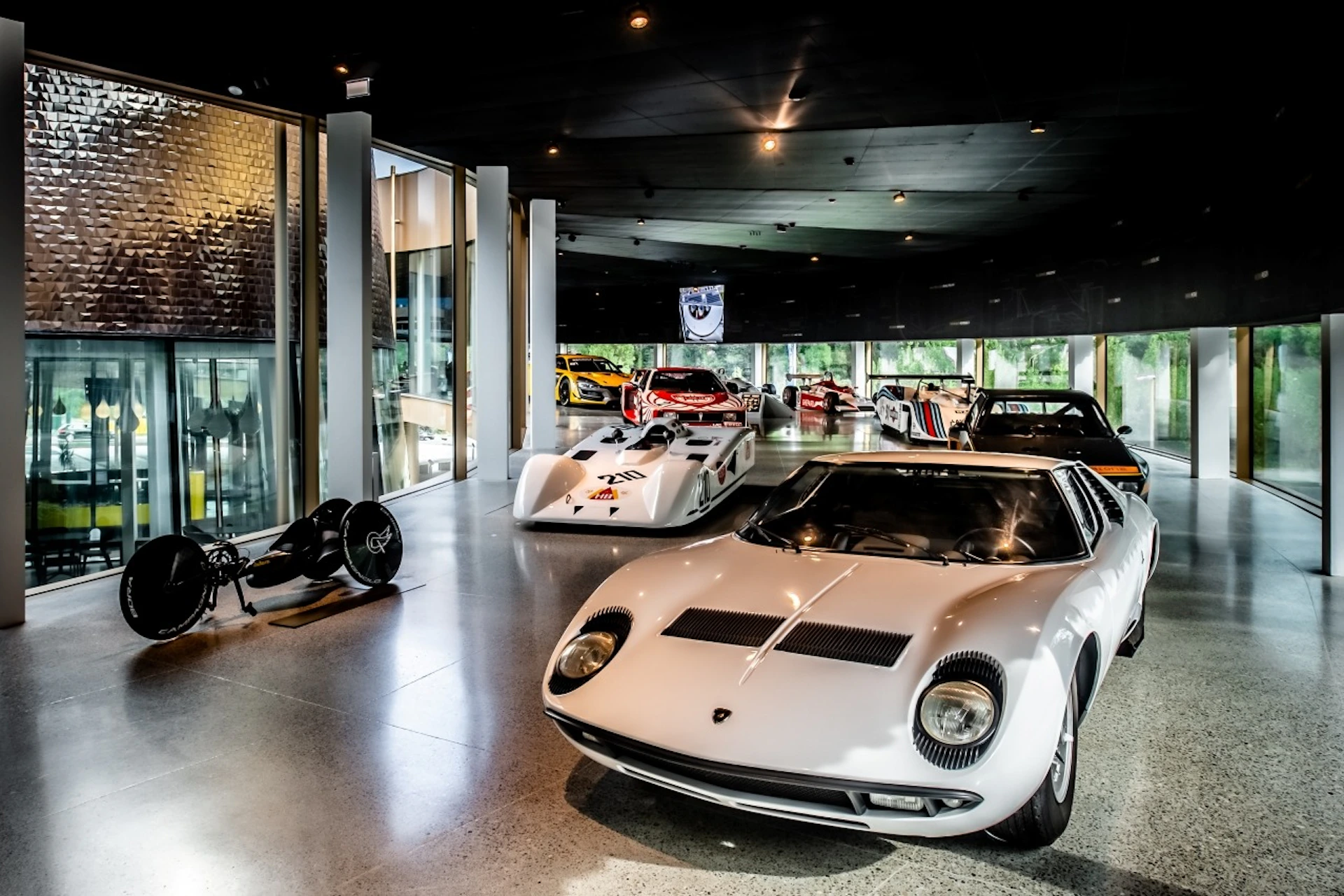 A collection of classic and modern racing cars displayed in a sleek, glass-walled showroom with reflective flooring.
