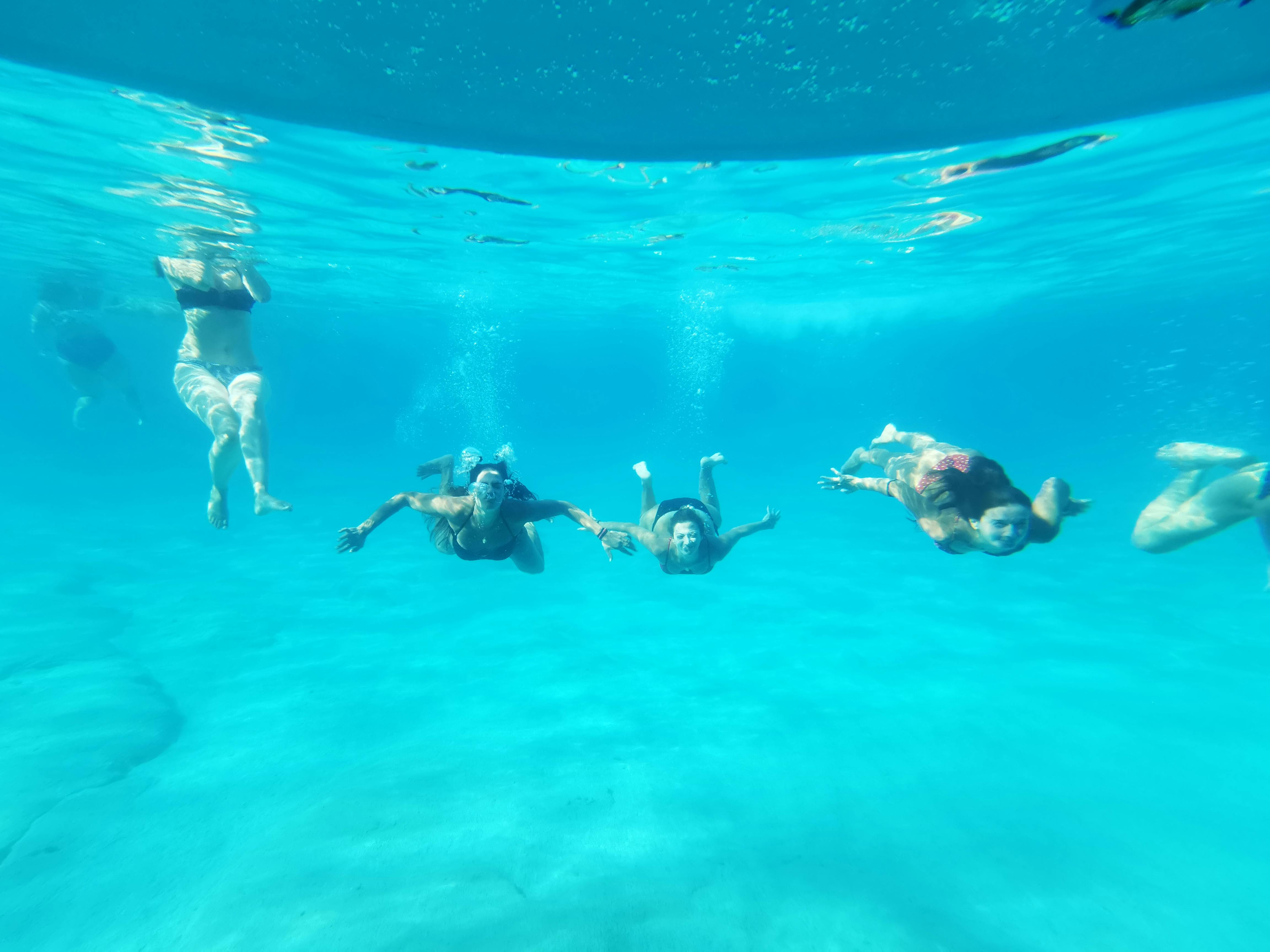 Τέσσερις άνθρωποι κολυμπούν κάτω από το νερό σε καταγάλανα νερά, που φαίνονται από κάτω, με το φως του ήλιου να αντανακλάται από πάνω.