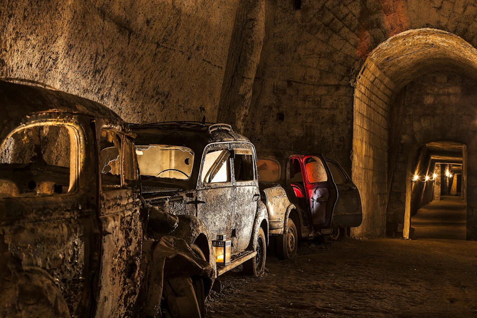 Three abandoned, rusted cars in a dimly lit, stone-walled tunnel, with one car door open and a lantern placed on the ground.