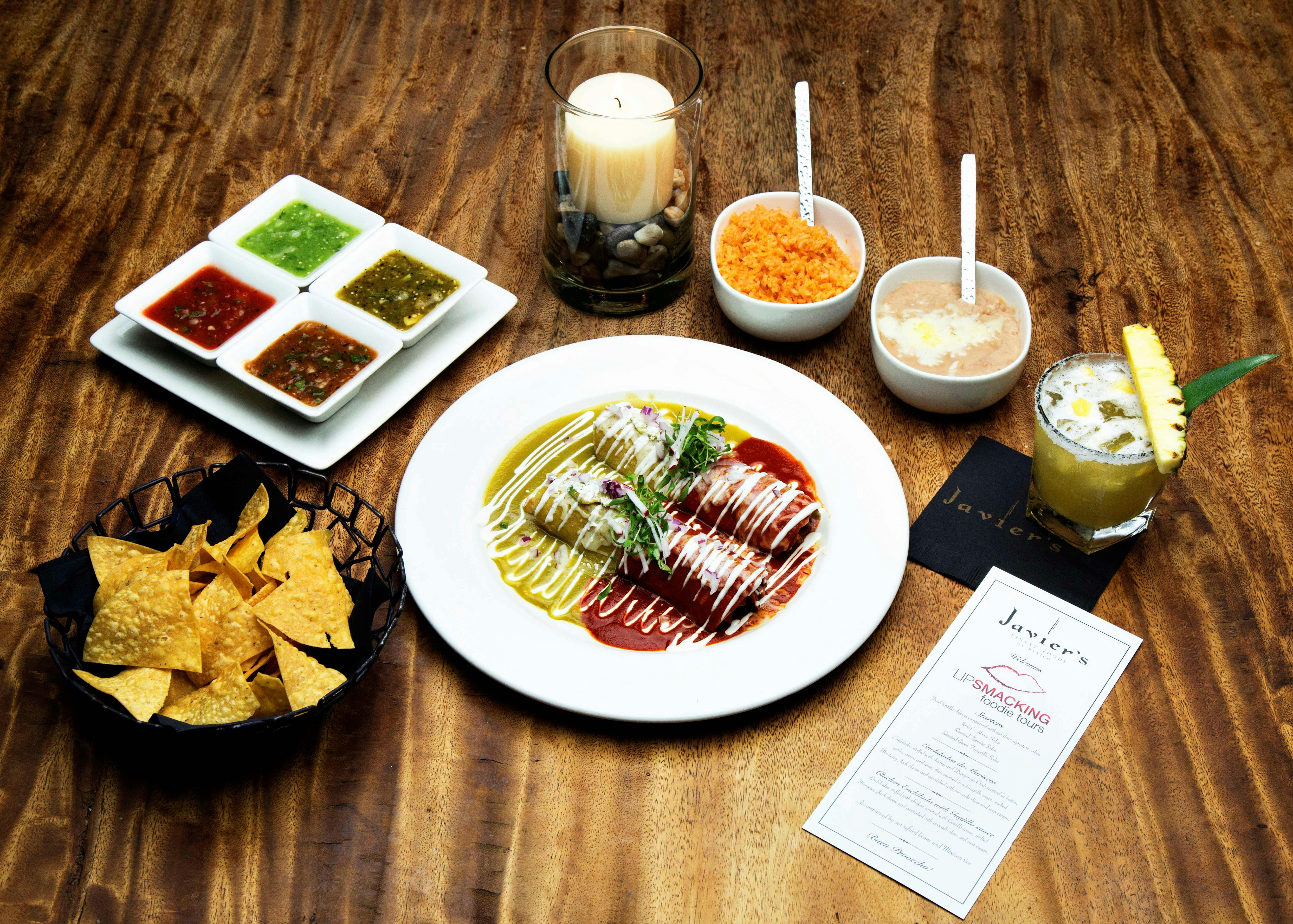 En tallerken med enchiladas med grønne og røde saucer, chips med dips, ris, bønner, et stearinlys og en drink på et træbord.