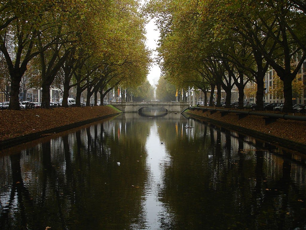 A serene canal flanked by trees with autumn foliage, leading to a stone bridge. Cars are parked along both sides of the water.