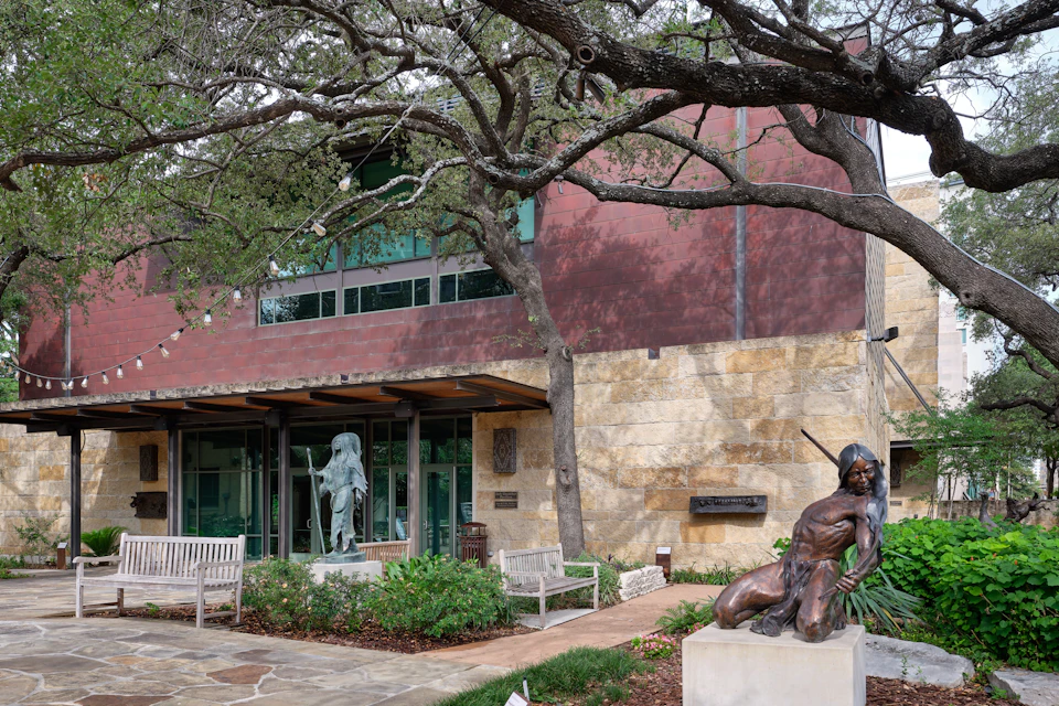 A small park with sculptures, benches, and trees in front of a modern building with stone and brick elements.