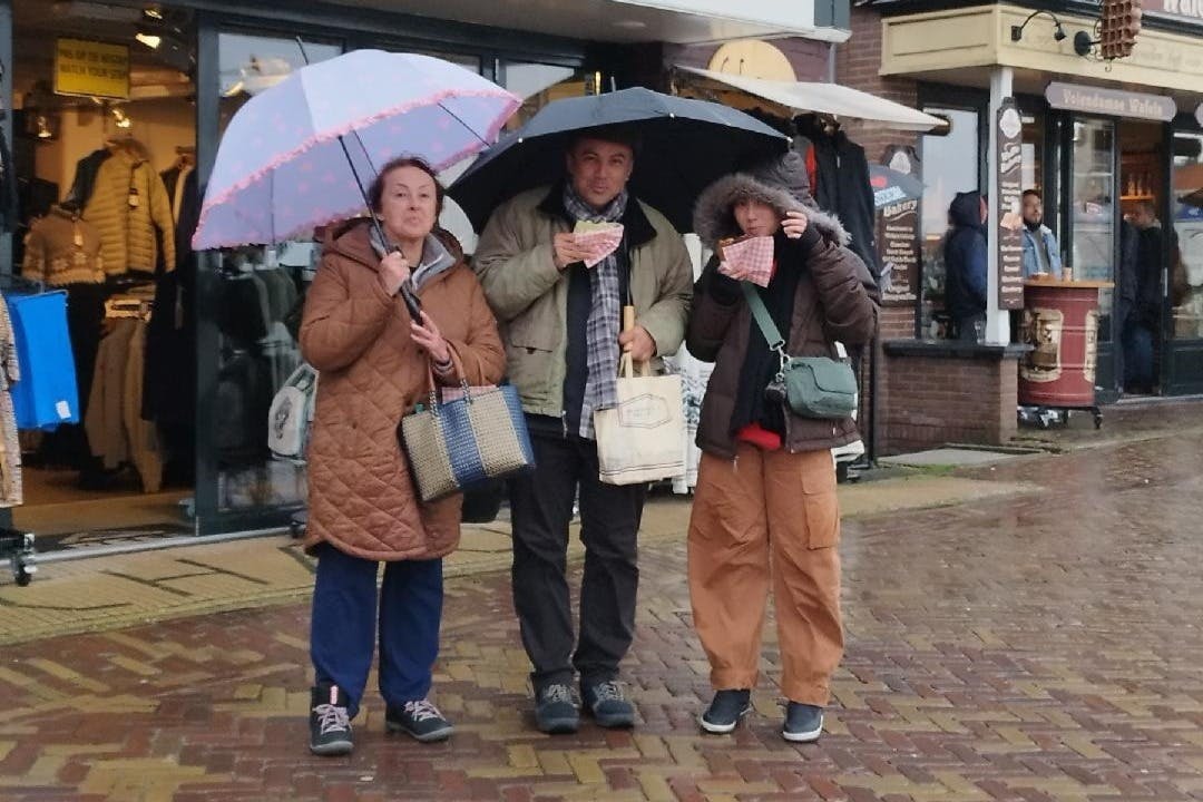 Τρία άτομα στέκονται κάτω από ομπρέλες μπροστά από ένα κατάστημα ρούχων σε ένα βρεγμένο πεζοδρόμιο.