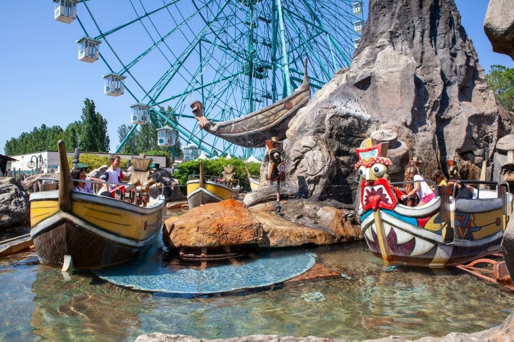Οι άνθρωποι οδηγούν θεματικές βάρκες σε ένα υδάτινο αξιοθέατο δίπλα σε τεχνητούς βράχους, με μια ρόδα λούνα παρκ στο παρασκήνιο.