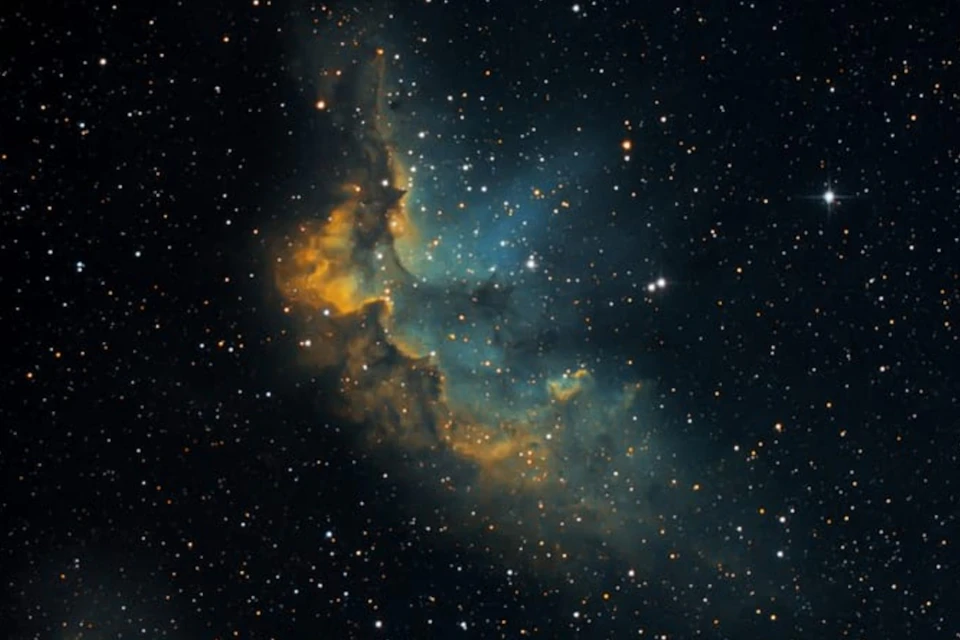 Colorful nebula in deep space with glowing blue, green, and orange gas clouds, surrounded by numerous stars.