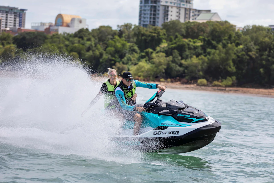 Two people riding a jet ski on a body of water with trees and urban buildings in the background.