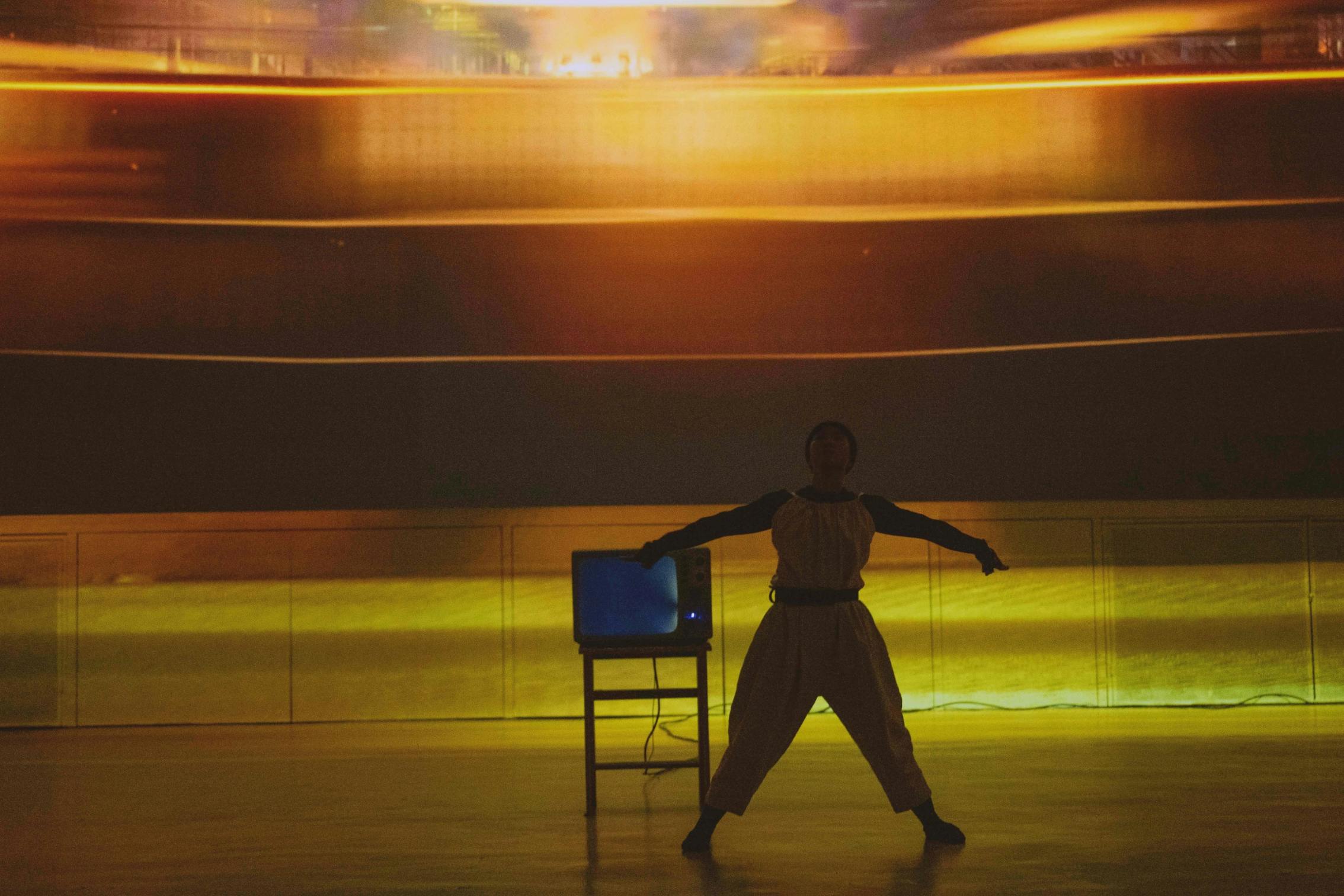 A person stands with arms extended near a small television on a stand, in front of a yellow-lit abstract background.