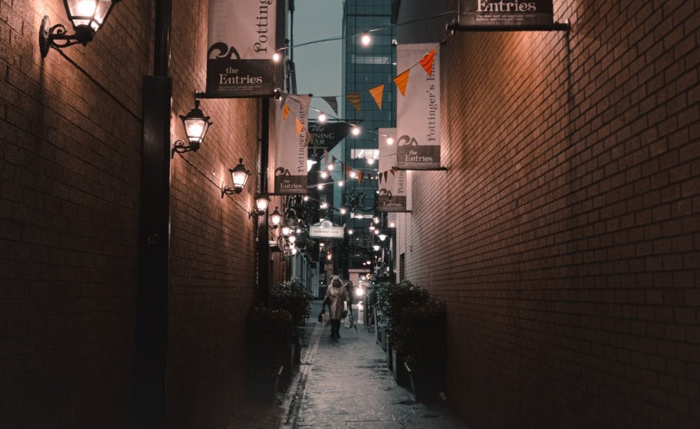 Un vicolo stretto con luci a corda, striscioni appesi, lanterne a muro e una persona che cammina verso la telecamera. Ambiente urbano al crepuscolo.