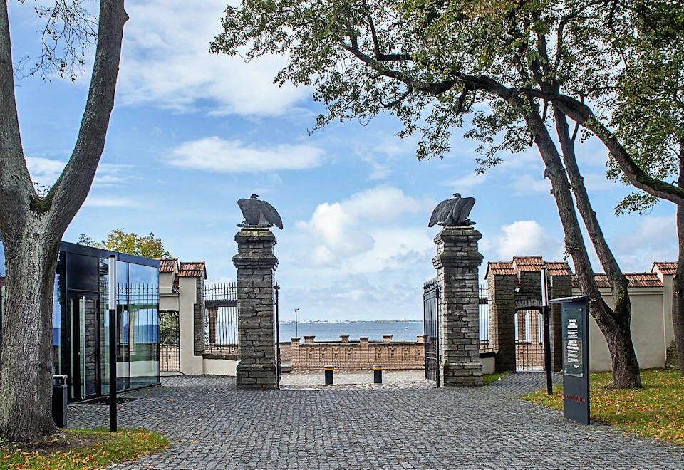 Pathway leading to a stone gate with eagle statues, flanked by trees, overlooking a body of water under a partly cloudy sky.