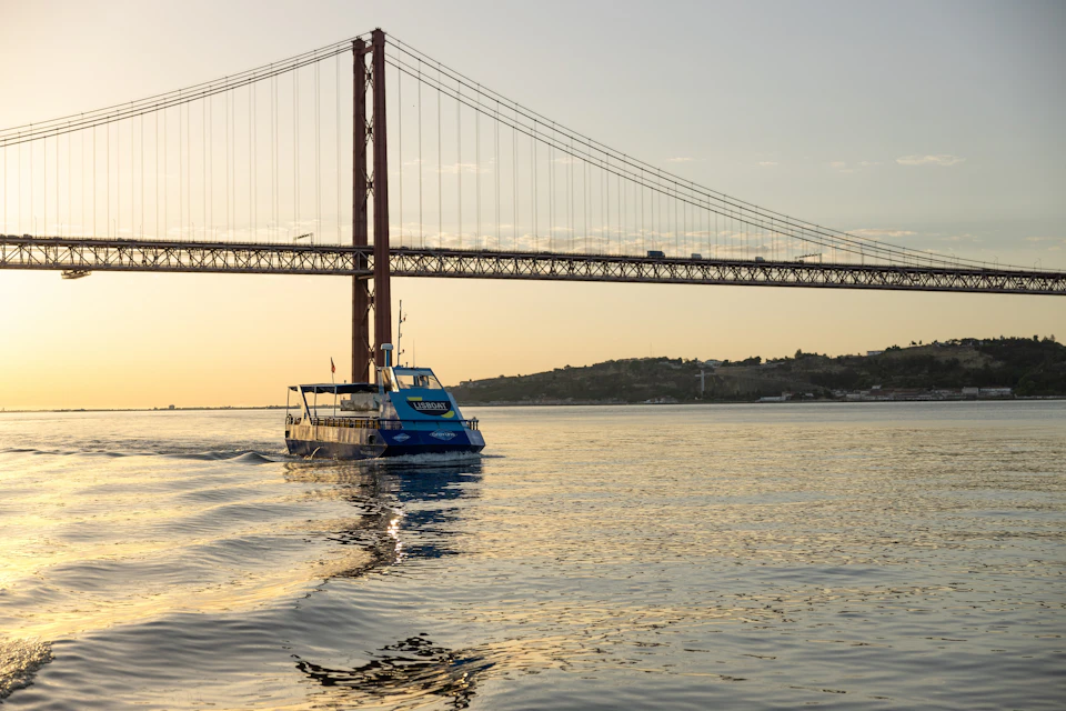 A blue boat cruising on calm waters with a large suspension bridge and hills in the background at sunset.