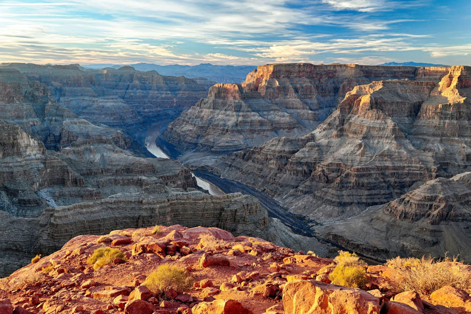 A view of the Grand Canyon with a river winding through the rocky landscape under a partly cloudy sky at sunset.