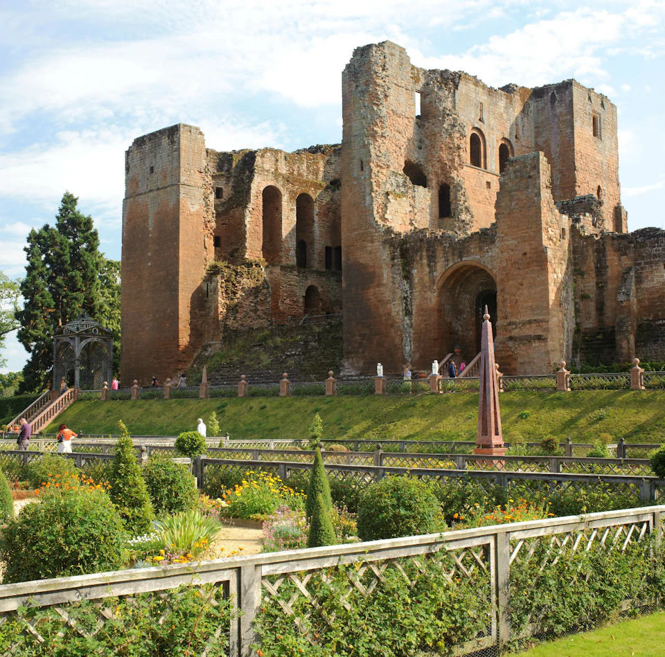 An ancient, partially ruined stone castle with arched windows, set behind a garden with manicured bushes and flowering plants.