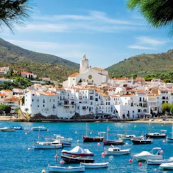 Seaside town with white buildings under a hilly landscape and blue sky. Boats are moored in calm harbor waters.