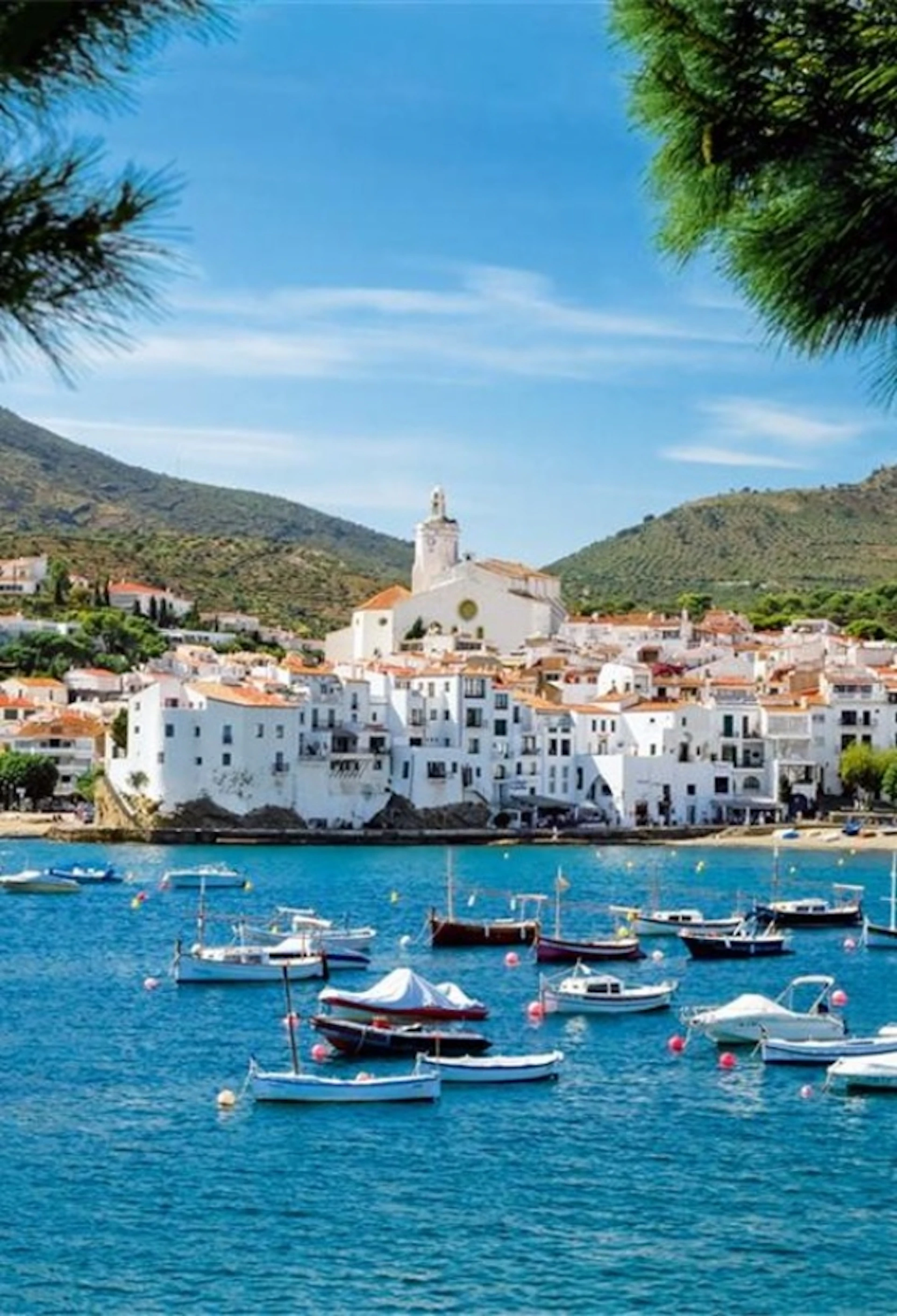 Seaside town with white buildings under a hilly landscape and blue sky. Boats are moored in calm harbor waters.