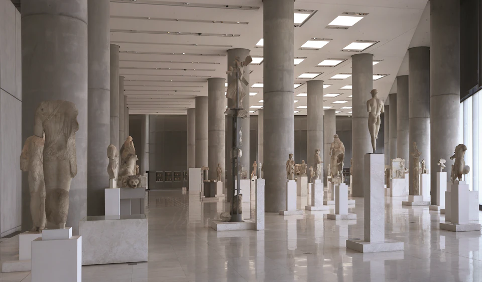 Spacious museum hall with rows of marble sculptures displayed on pedestals, surrounded by large columns under a bright ceiling.