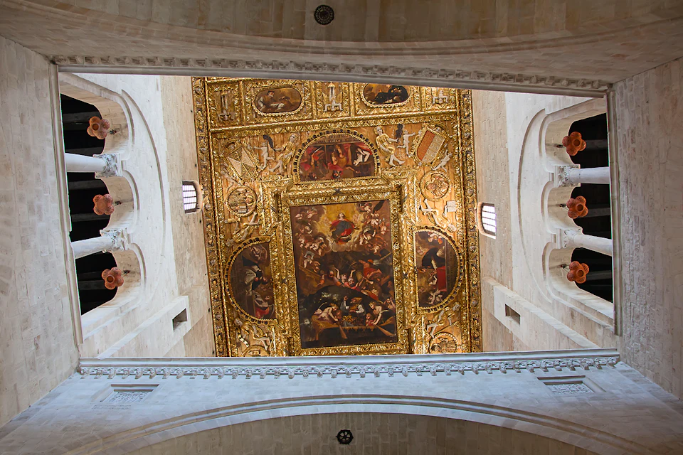 Ceiling of a historic building with intricate gold decorations and religious paintings, surrounded by arched stone walls.