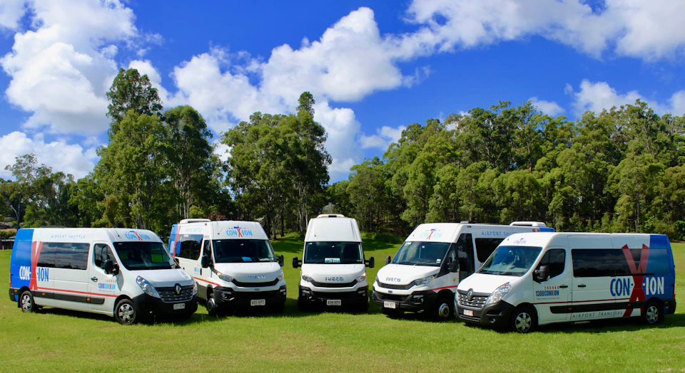 A row of white vans parked on grass in front of a forested area under a bright blue sky with scattered clouds.