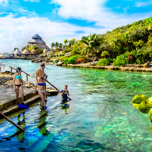 Menschen in Badekleidung und mit Schnorchel, die sich darauf vorbereiten, eine klare, tropische Lagune zu betreten, die von üppigem Grün und strohgedeckten Gebäuden umgeben ist.