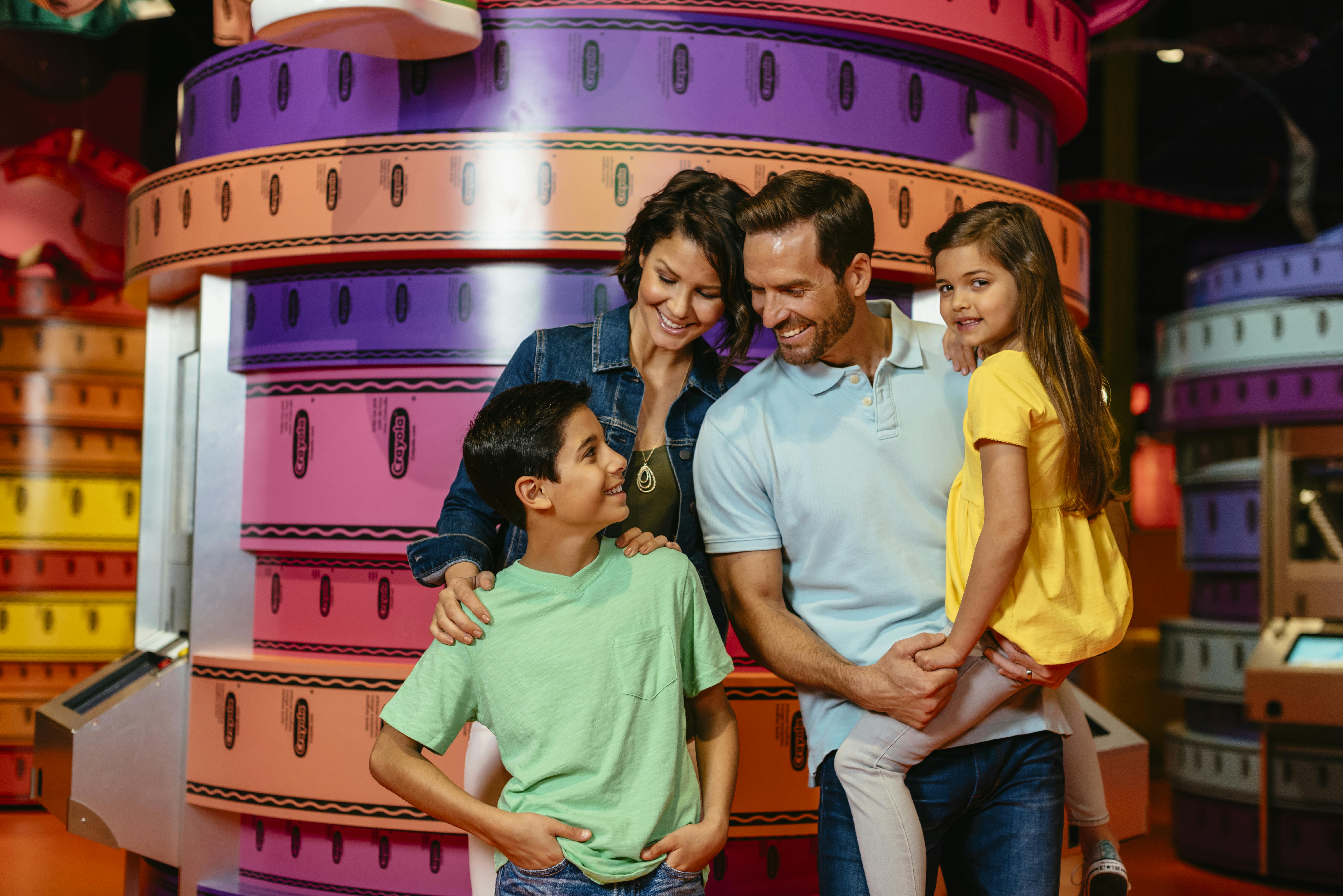 円形のオブジェクトや装飾が積み重なったカラフルな背景の前には、笑顔の4人家族が立っています。