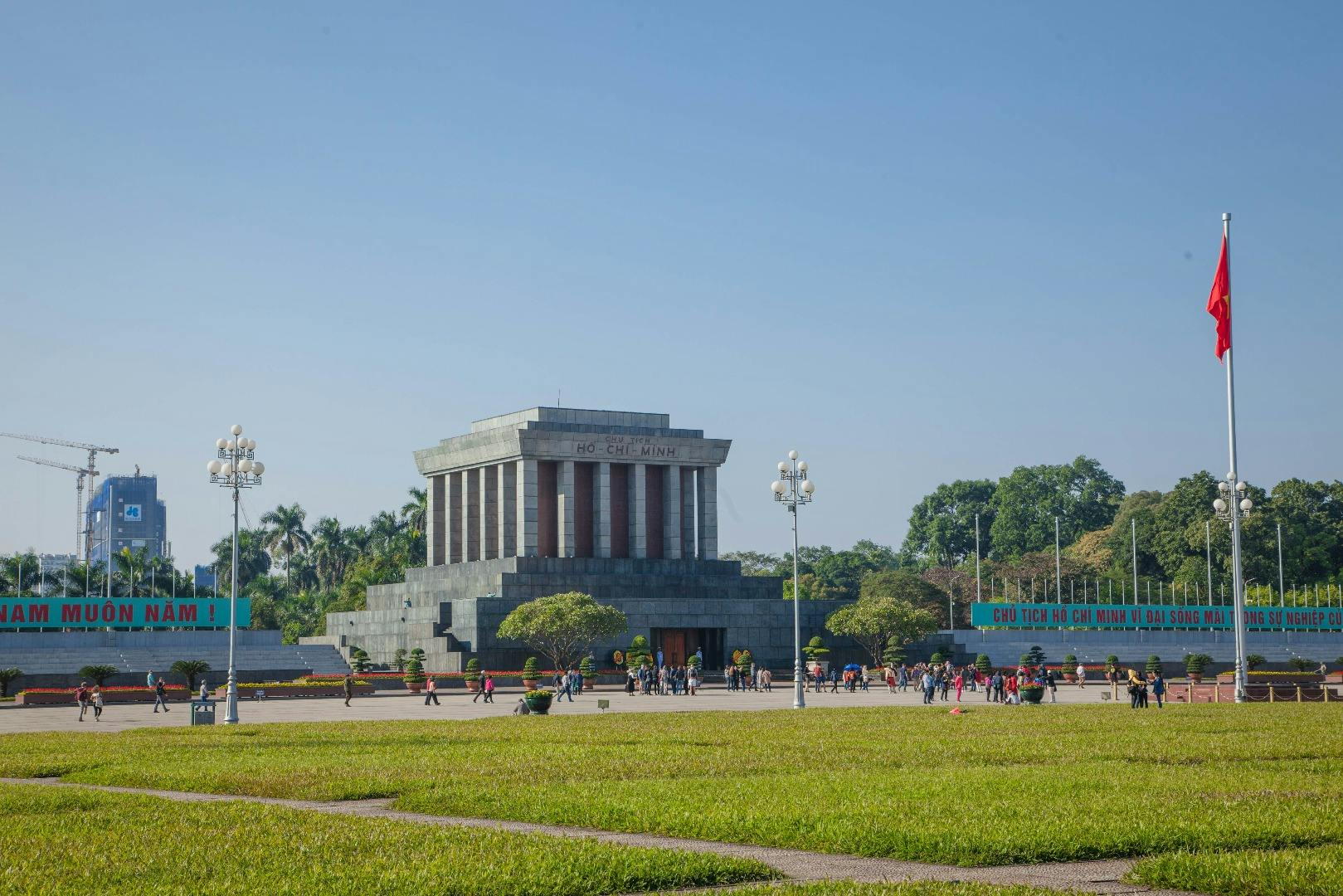 Duży kamienny budynek z napisem „Ho Chi Minh”, otoczony zielenią, ludźmi i czerwoną flagą na pierwszym planie.